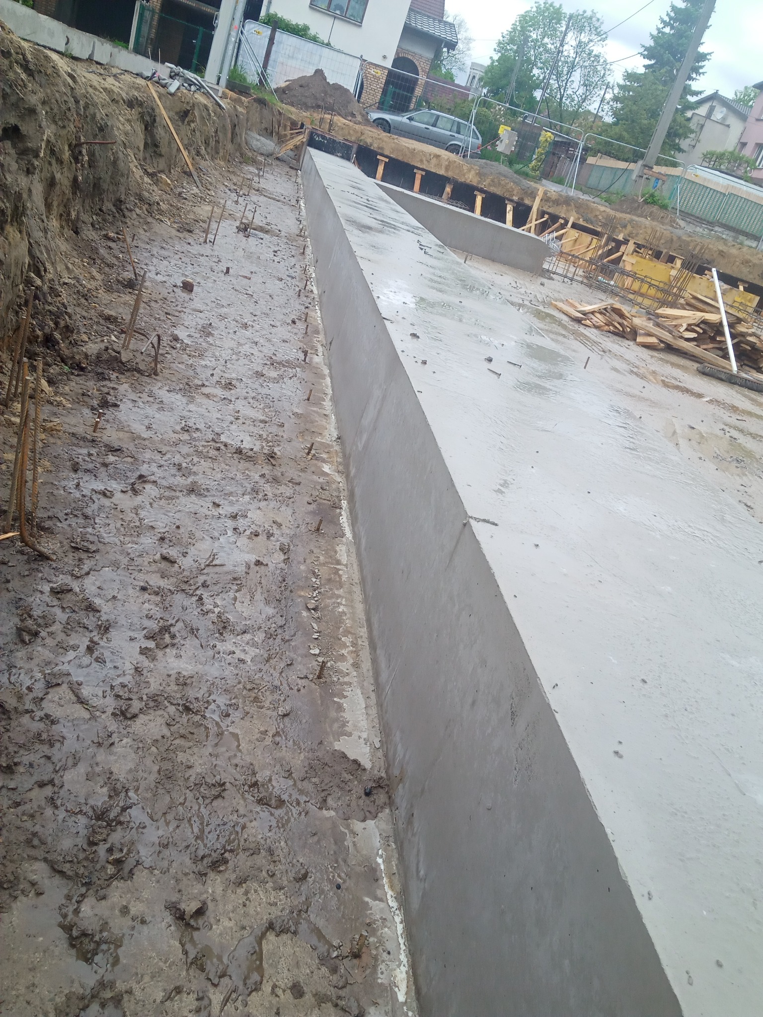 Świeżo wylany, mokry betonowy fundament w wykopie, widoczne wystające pręty zbrojeniowe i drewniane szalunki, w tle zabudowa jednorodzinna i zaparkowany samochód.