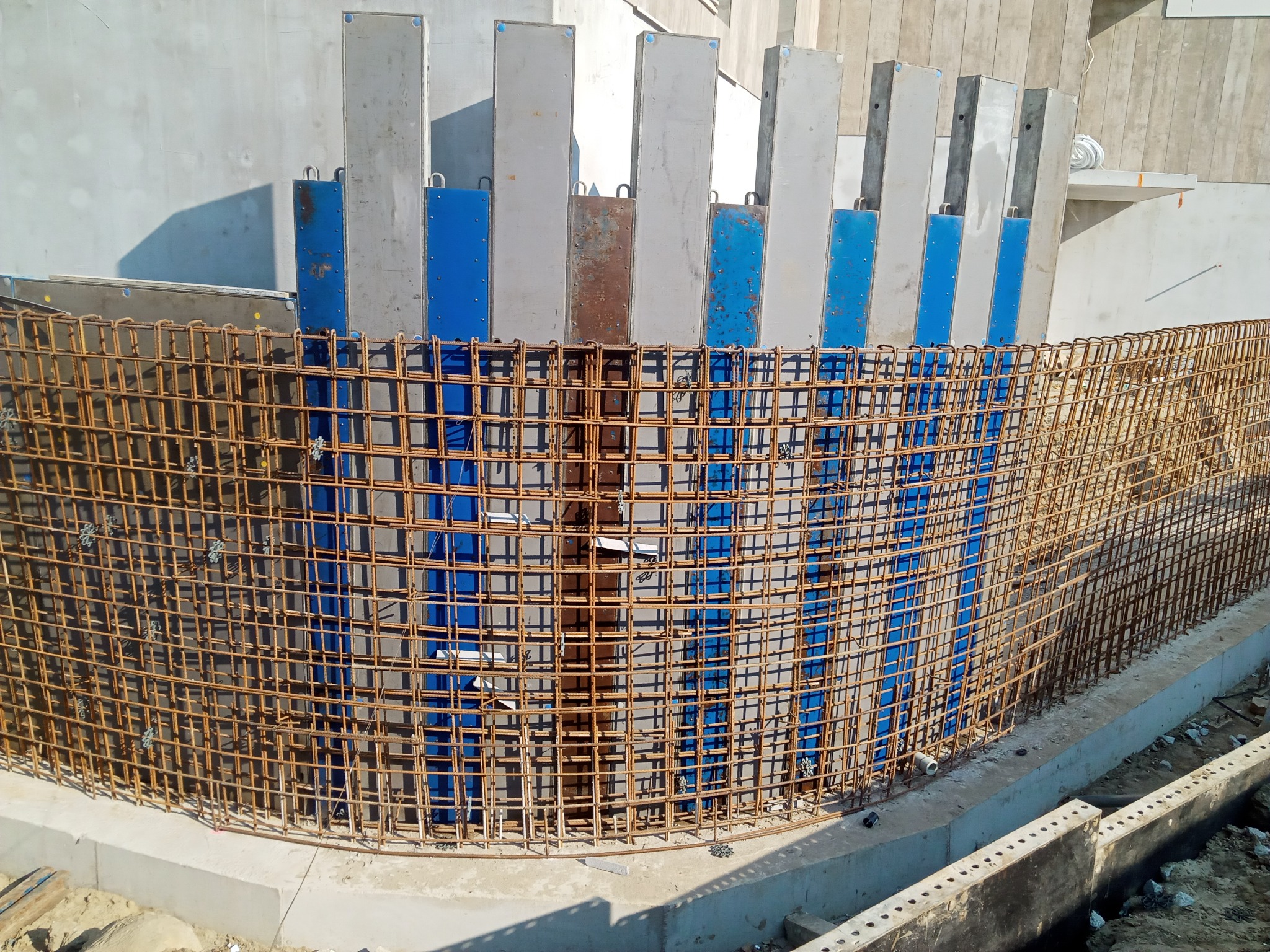 Zbrojenie łukowe fundamentu z szalunkiem stalowym w kolorze niebieskim, widoczne spinki montażowe, betonowa podstawa i ściana w tle.