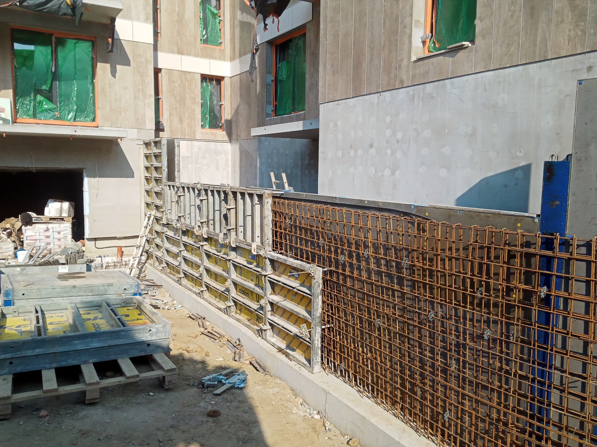 Zbrojenie pionowe fundamentu z widocznymi elementami szalunkowymi i konstrukcją budynku w tle.