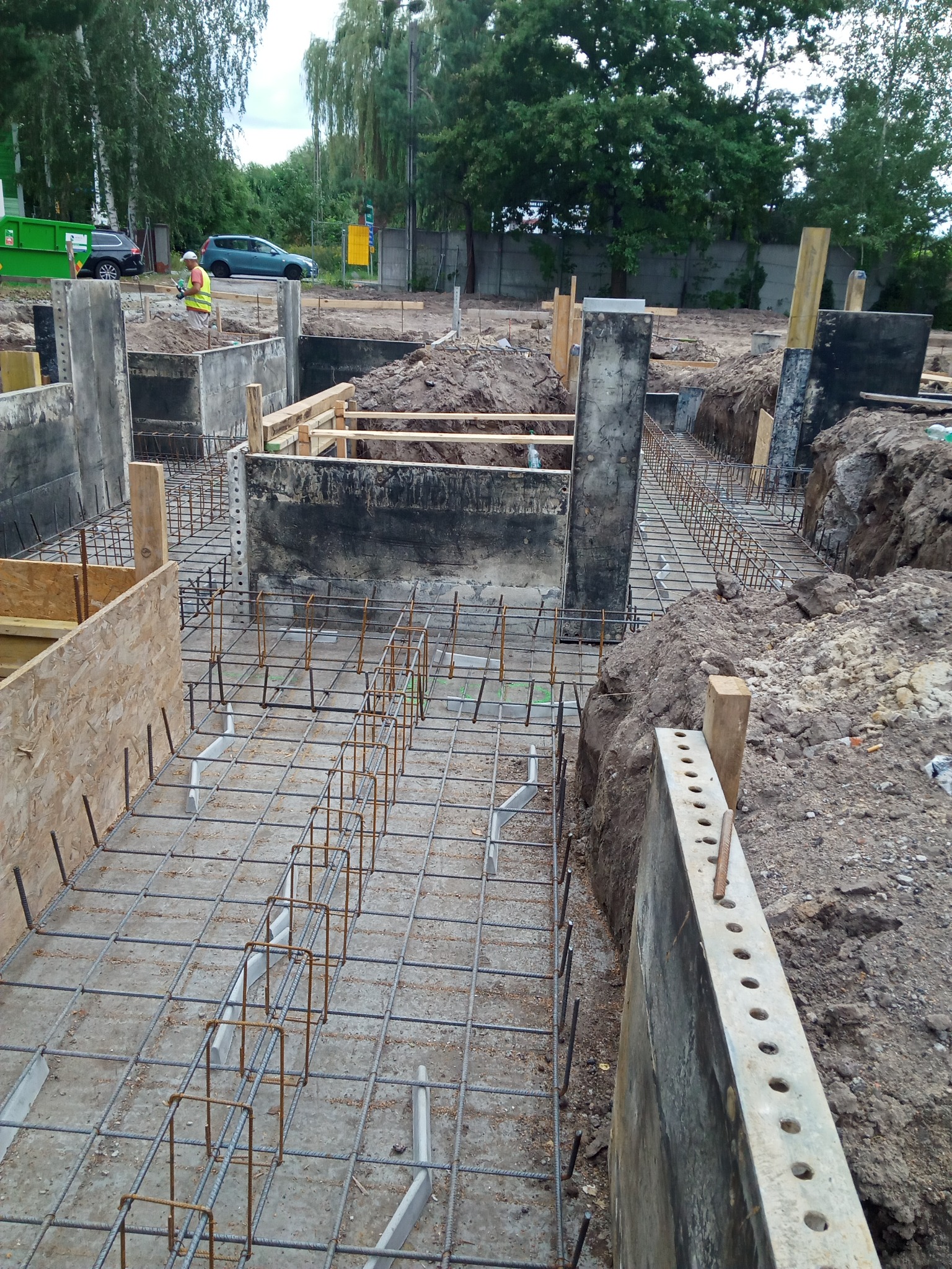 Zbrojenie fundamentów budynku w trakcie budowy, widoczne betonowe szalunki, stalowe pręty zbrojeniowe, wykopane rowy i pracownik w oddali.