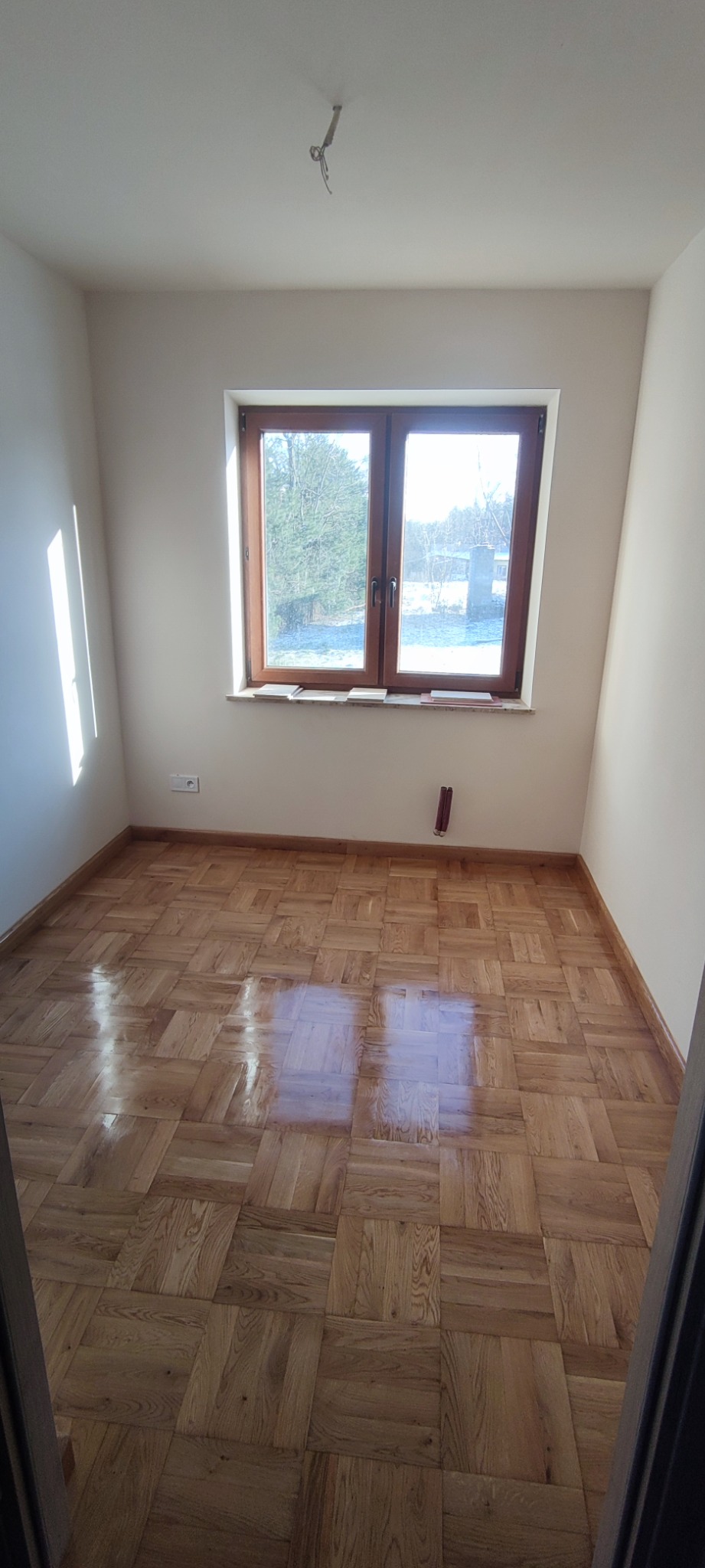 Pusty pokój po remoncie z parkietem ułożonym w kwadraty, świeżo pomalowanymi ścianami na biało i widokiem na zalesiony teren za oknem.