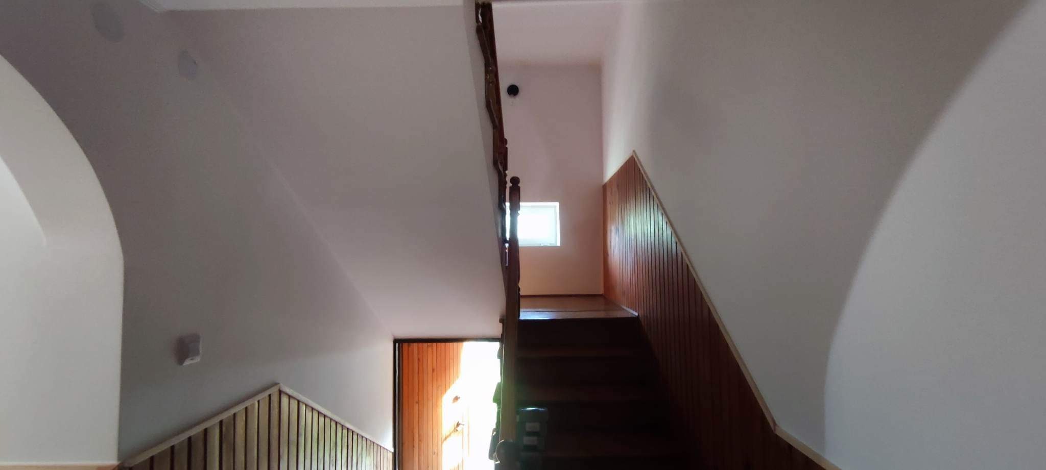 Klatka schodowa z drewnianymi schodami i boazerią, świeżo pomalowane ściany na biało, w tle widoczne okno.