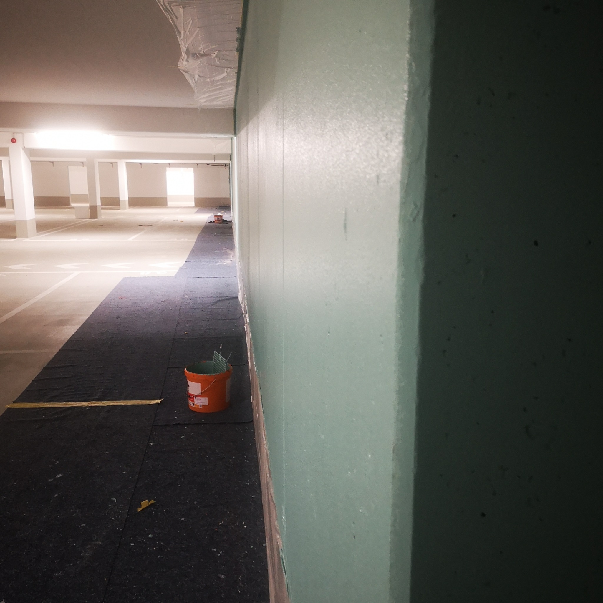Świeżo pomalowana, jasnozielona ściana w garażu podziemnym, zabezpieczona czarną folią i pomarańczowym wiadrem z farbą u podstawy.
