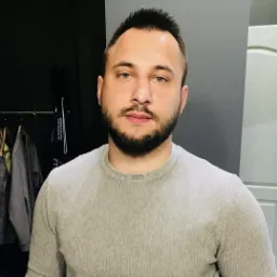 Portret mężczyzny z brodą w szarym swetrze na tle szarej ściany z elementami garderoby w tle.