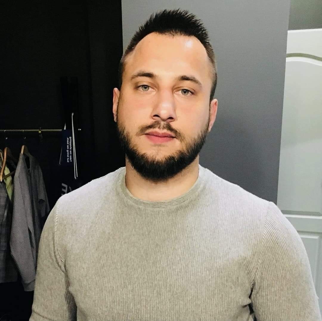 Portret mężczyzny z brodą w szarym swetrze na tle szarej ściany z elementami garderoby w tle.