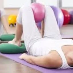 Kobieta leżąca na plecach na fioletowej macie, z piłką do ćwiczeń między kolanami, stopy oparte na zielonych poduszkach, widok z boku, w tle lustro odbijające ćwiczącą osobę.