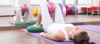 Kobieta leżąca na plecach na fioletowej macie, z piłką do ćwiczeń między kolanami, stopy oparte na zielonych poduszkach, widok z boku, w tle lustro odbijające ćwiczącą osobę.