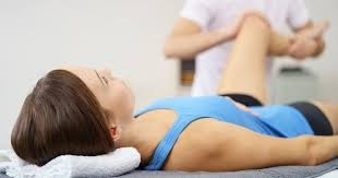 Pacjentka leżąca na plecach podczas badania kolana przez terapeutę w białym uniformie, jasne otoczenie gabinetu.