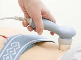 Ultradźwięki aplikowane na dolną część pleców pacjenta za pomocą głowicy z szarym uchwytem i niebieskim pasem mocującym elektrodę.