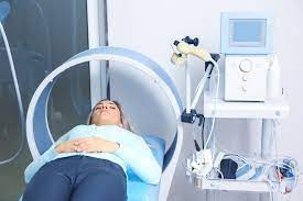 Pacjentka leżąca pod aparaturą do terapii, widoczne elektrody i panel sterujący, jasne wnętrze gabinetu.