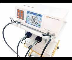 Białe urządzenie medyczne z wyświetlaczem pokazującym parametry zabiegu, podłączone przewodami zakończonymi czarnymi elektrodami. Widoczne przyciski sterujące i logo producenta.