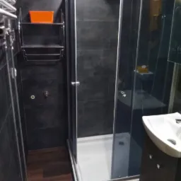 Nowoczesna łazienka z ciemnymi płytkami, kabiną prysznicową z przesuwnymi drzwiami, drewnopodobną podłogą i chromowanym grzejnikiem drabinkowym z pomarańczowym pojemnikiem na półce.