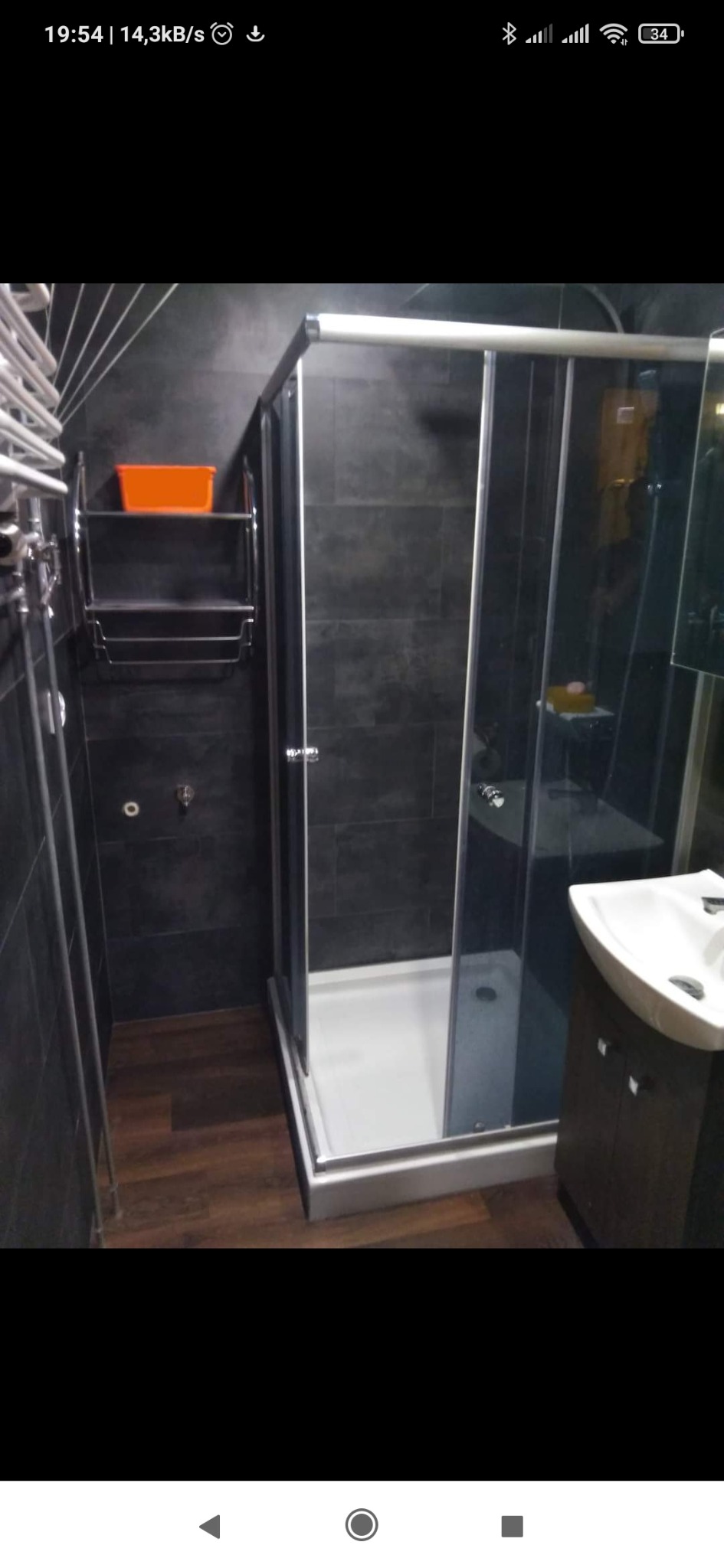 Nowoczesna łazienka z ciemnymi płytkami, kabiną prysznicową z przesuwnymi drzwiami, drewnopodobną podłogą i chromowanym grzejnikiem drabinkowym z pomarańczowym pojemnikiem na półce.