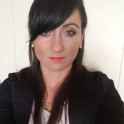 Portret kobiety z ciemnymi włosami, w czarnej marynarce i białej bluzce, z delikatnym makijażem i kolczykami w kształcie serc.