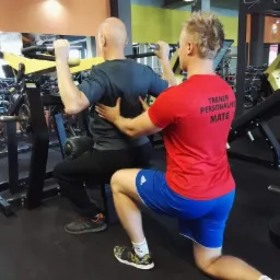 Mężczyzna ćwiczy na maszynie pod nadzorem instruktora w czerwonej koszulce z napisem 'TRENER PERSONALNY MATE' w siłowni.