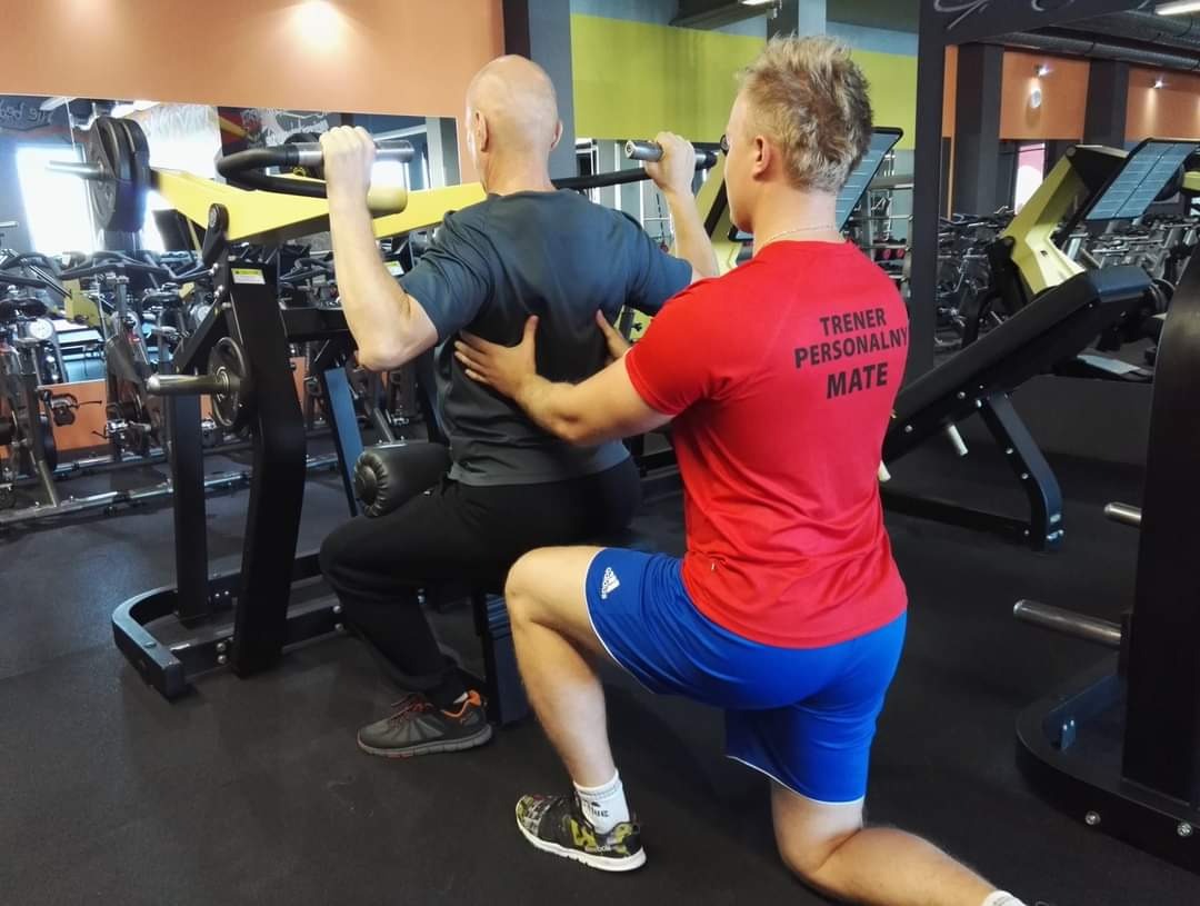 Mężczyzna ćwiczy na maszynie pod nadzorem instruktora w czerwonej koszulce z napisem 'TRENER PERSONALNY MATE' w siłowni.