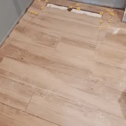 Układanie podłogi z płytek imitujących drewno, z użyciem ż&oacute;łtych krzyżyk&oacute;w dystansowych, widoczne niedociągnięcia przy ścianie i pozostałości fugi.