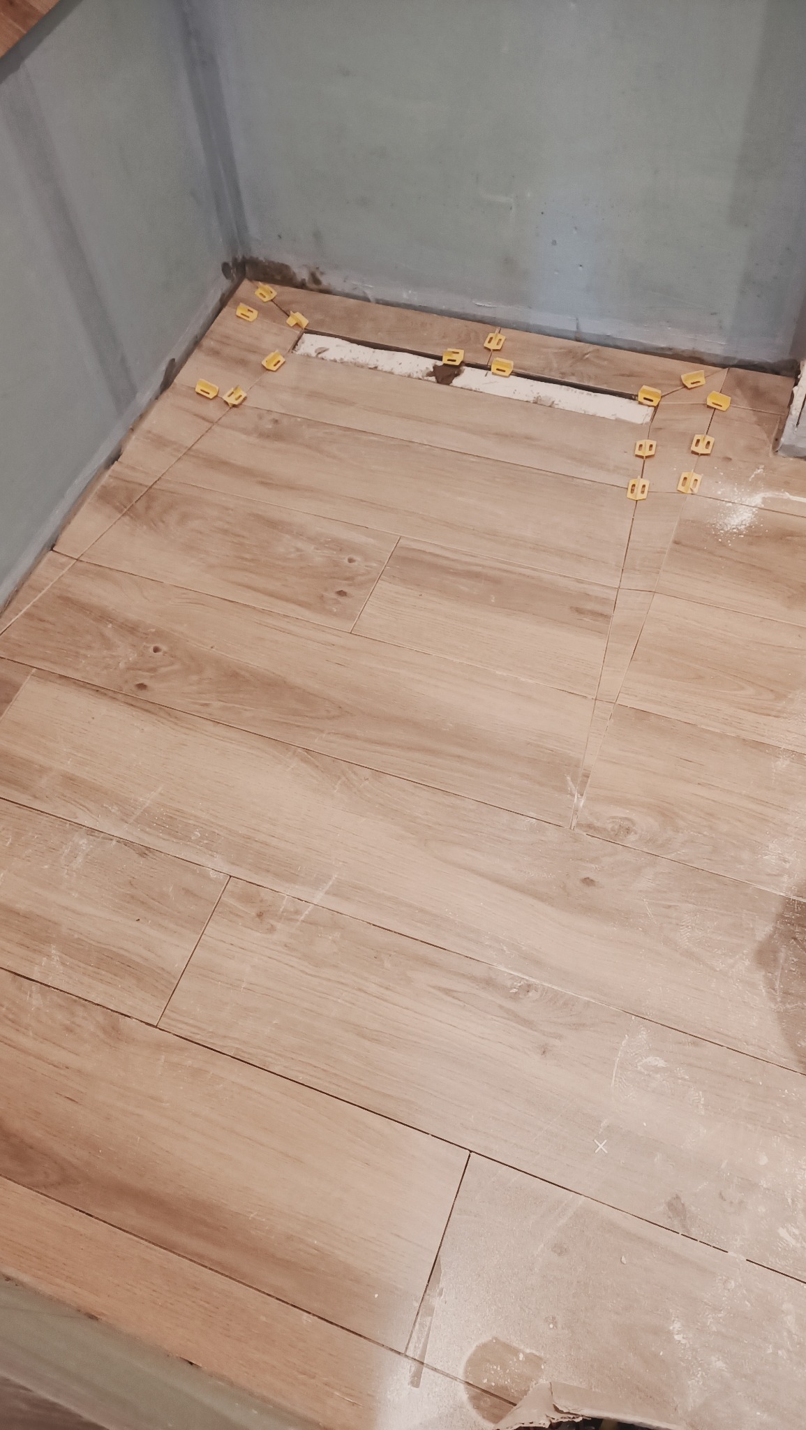 Układanie podłogi z płytek imitujących drewno, z użyciem żółtych krzyżyków dystansowych, widoczne niedociągnięcia przy ścianie i pozostałości fugi.