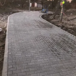 Świeżo ułożona, zakrzywiona ścieżka z szarych kostek brukowych w trakcie prac wykończeniowych; w tle widoczny słup energetyczny i wózek transportowy.