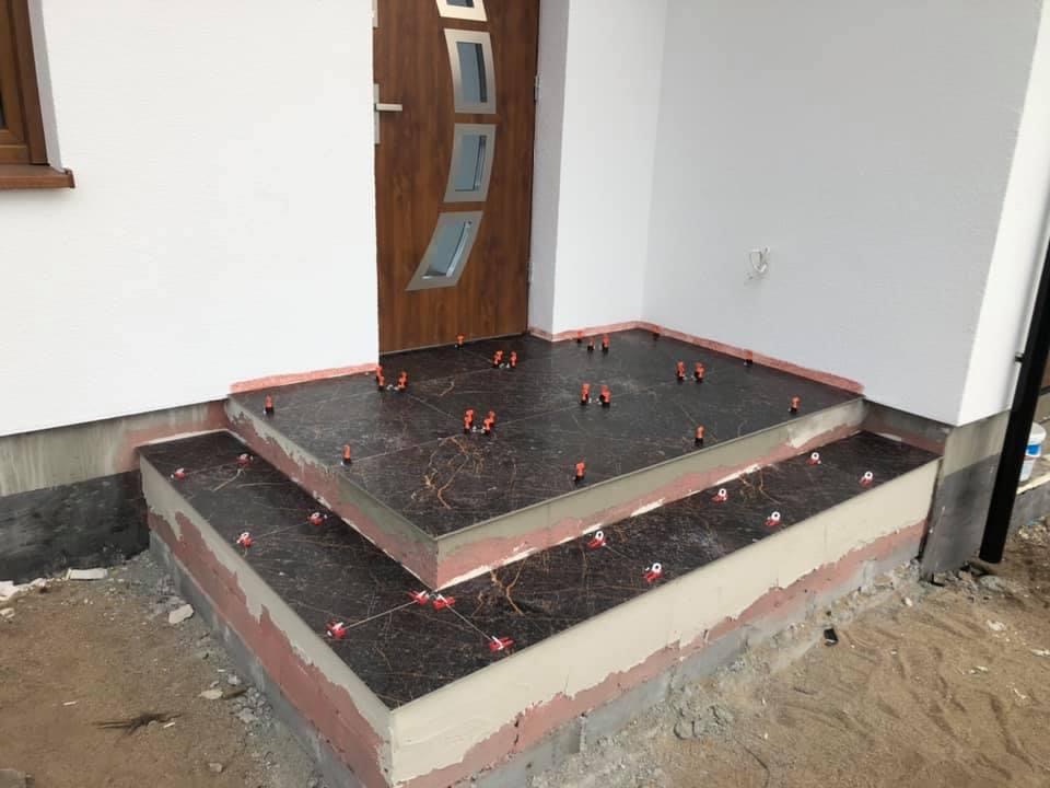 Kładzenie płytek imitujących marmur na schodach wejściowych, z użyciem systemu poziomowania płytek (klipsy i kliny), widoczne ślady zaprawy, biała elewacja i brązowe drzwi.