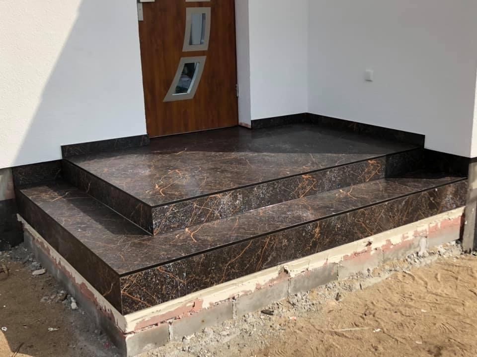 Wykończone płytkami schody wejściowe z ciemnego marmuru z pomarańczowymi żyłkami, prowadzące do drewnianych drzwi z geometrycznymi przeszkleniami, widoczne niedokończone prace na fundamencie.