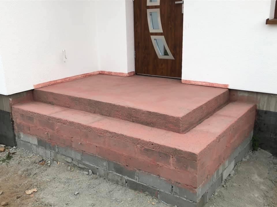 Dwustopniowe schody wejściowe do domu, pomalowane na kolor ceglasty, z widoczną białą elewacją i brązowymi drzwiami z przeszkleniami.