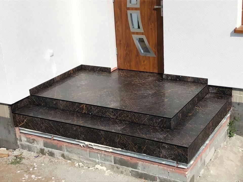 Wykończone marmurowe schody wejściowe do domu, z widoczną drewnianą stolarką drzwiową i fragmentem otynkowanej elewacji.