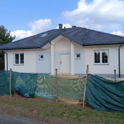 Nowoczesny dom jednorodzinny z szarą dachówką i elementami wykończenia elewacji w odcieniach bieli i szarości, częściowo zasłonięty zieloną siatką budowlaną.