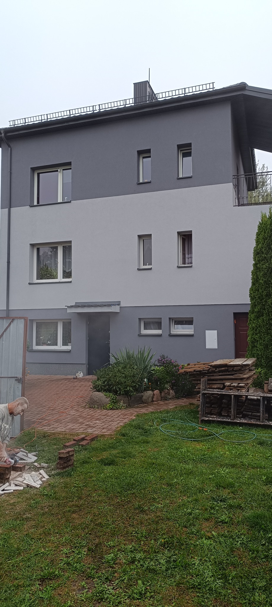 Dwukondygnacyjny dom z elewacją w odcieniach szarości, widok z ogrodu, z pracującym mężczyzną przy układaniu kostki brukowej na podjeździe.