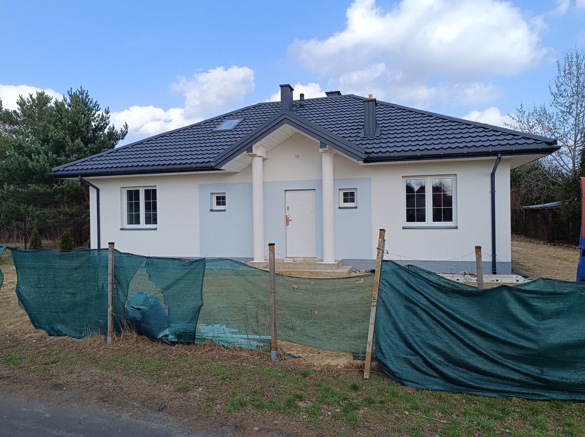 Nowoczesny dom jednorodzinny z szarą dachówką i elementami wykończenia elewacji w odcieniach bieli i szarości, częściowo zasłonięty zieloną siatką budowlaną.