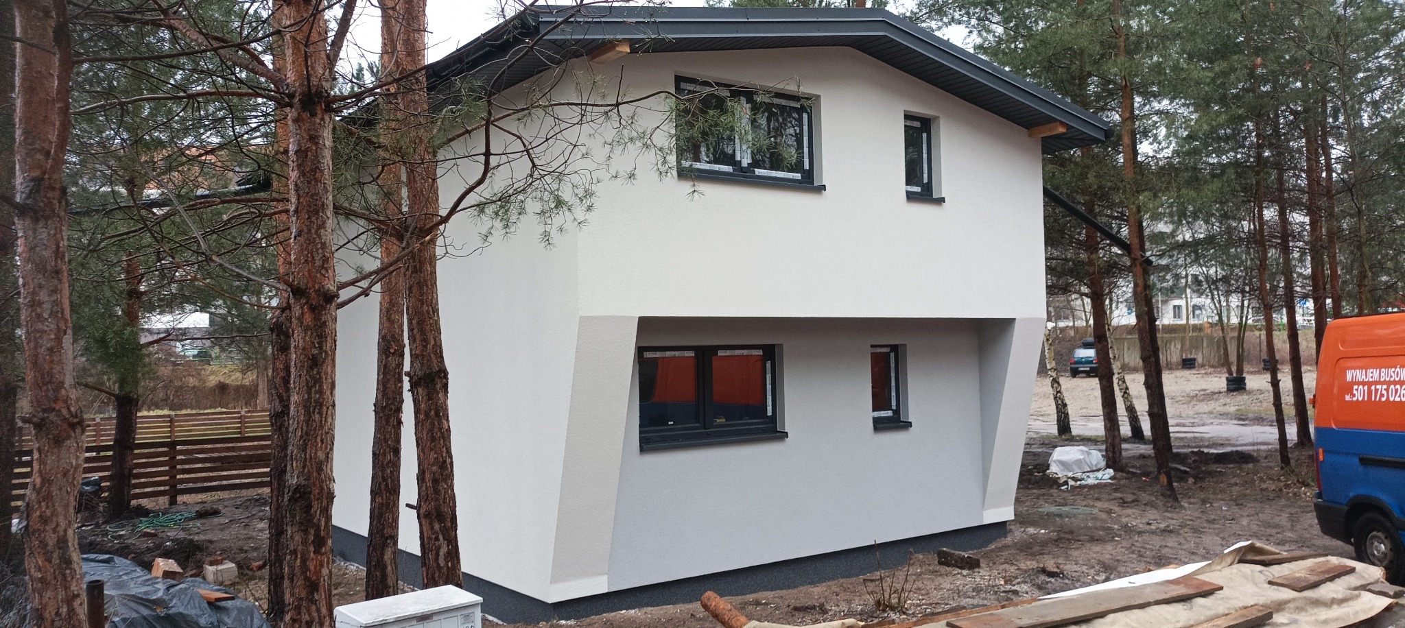 Dwukondygnacyjny dom z jasną elewacją, widoczny z perspektywy ogrodu, z elementami nowoczesnej architektury w postaci skośnych ścian i czarnych obramowań okiennych, otoczony drzewami iglastymi.