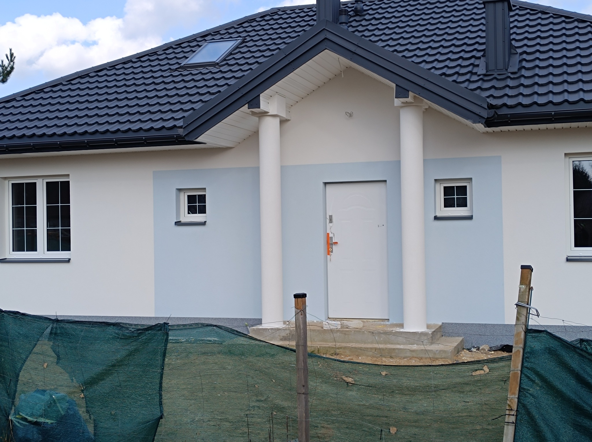 Nowoczesny dom jednorodzinny z jasną elewacją, grafitowym dachem i kolumnami przy wejściu, zabezpieczony siatką ochronną w trakcie prac wykończeniowych.