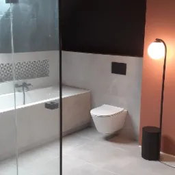 Nowoczesna łazienka z kabiną prysznicową typu walk-in, podwieszaną toaletą, białą wanną z czarną armaturą i oryginalną mozaiką nad wanną, oświetlona lampą podłogową z kulistym kloszem.