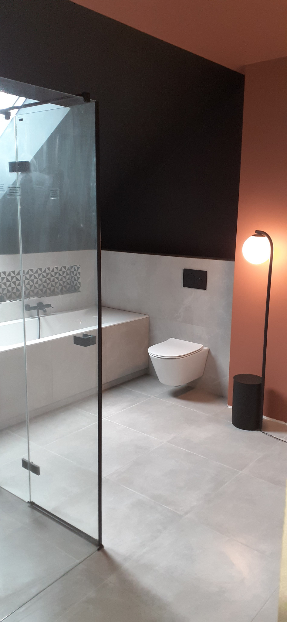 Nowoczesna łazienka z kabiną prysznicową typu walk-in, podwieszaną toaletą, białą wanną z czarną armaturą i oryginalną mozaiką nad wanną, oświetlona lampą podłogową z kulistym kloszem.