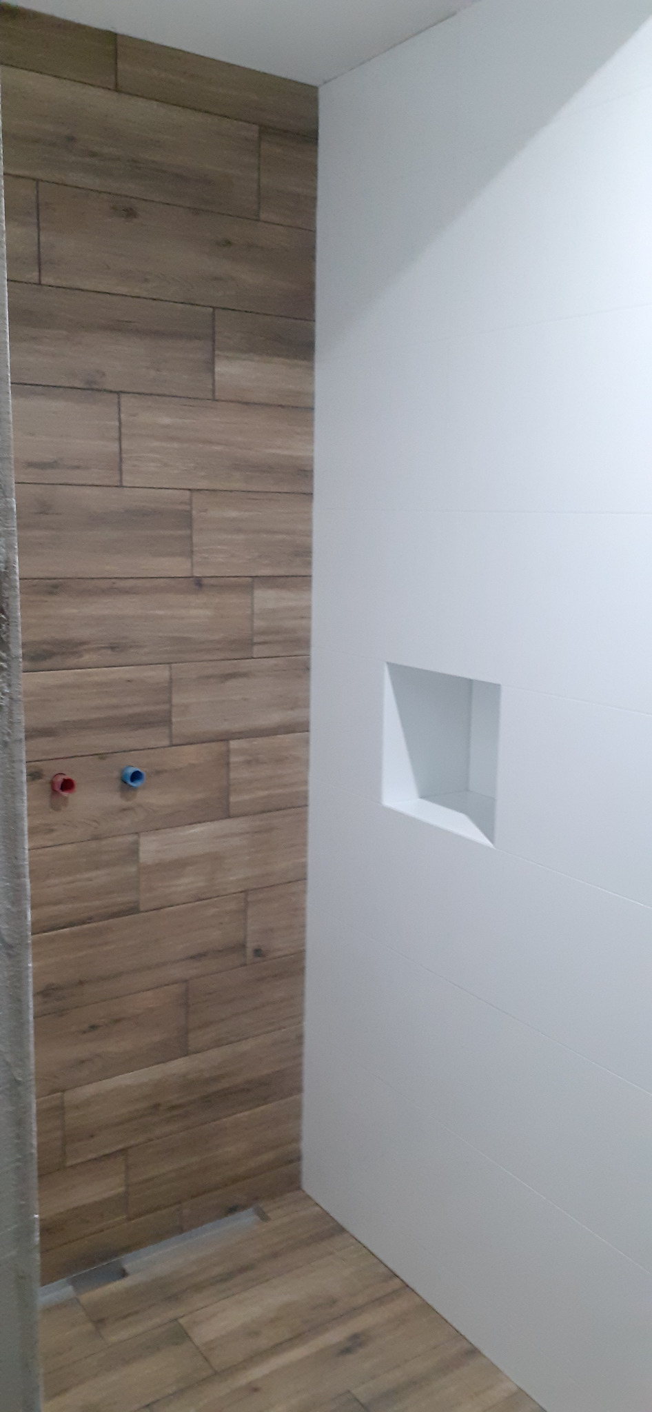 Wykończona ściana łazienki z drewnopodobnymi płytkami, widoczne przyłącza wodne z niebieską i czerwoną zaślepką oraz biała ściana z wnęką.