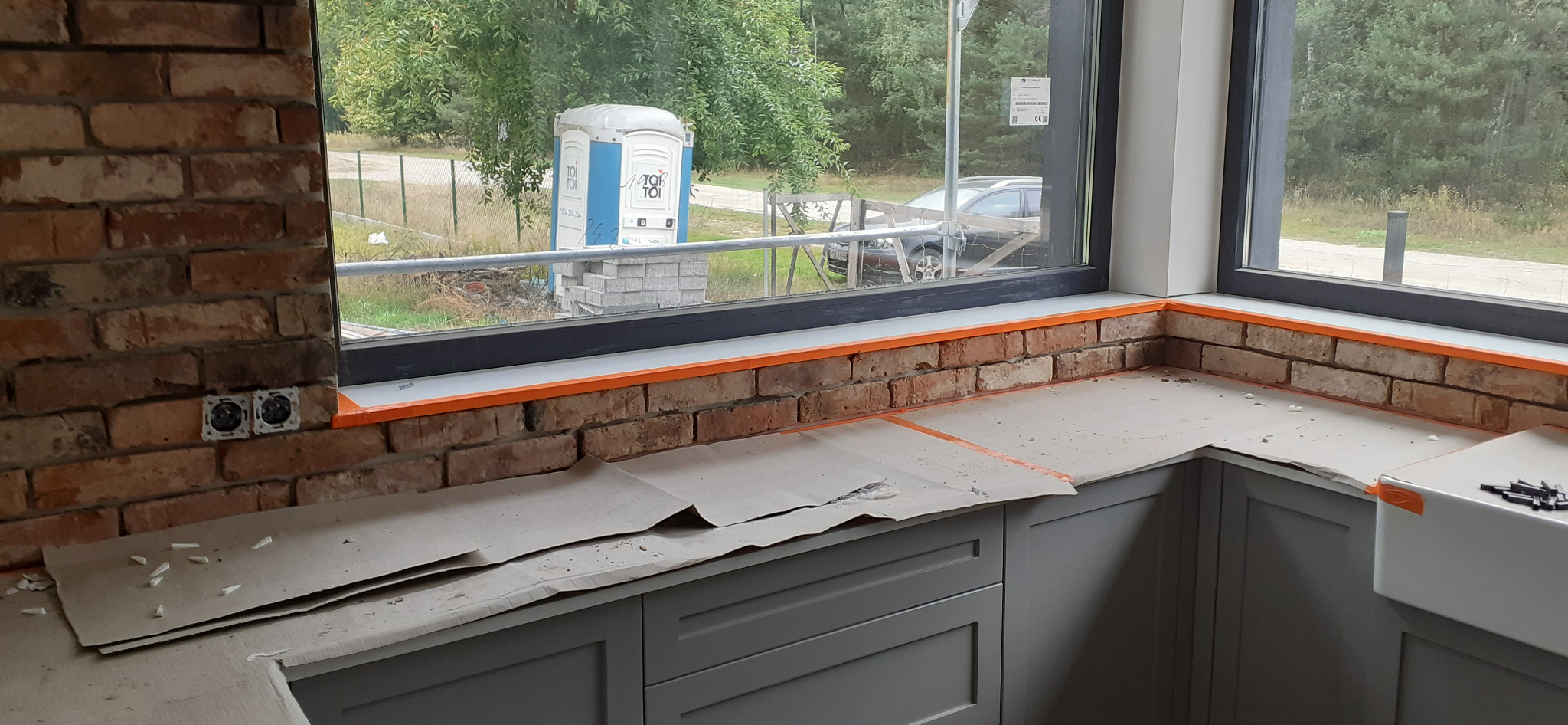 Kuchnia w trakcie wykańczania z ceglaną ścianą, zabezpieczonym blatem, szarą zabudową i widokiem na przenośną toaletę na zewnątrz.