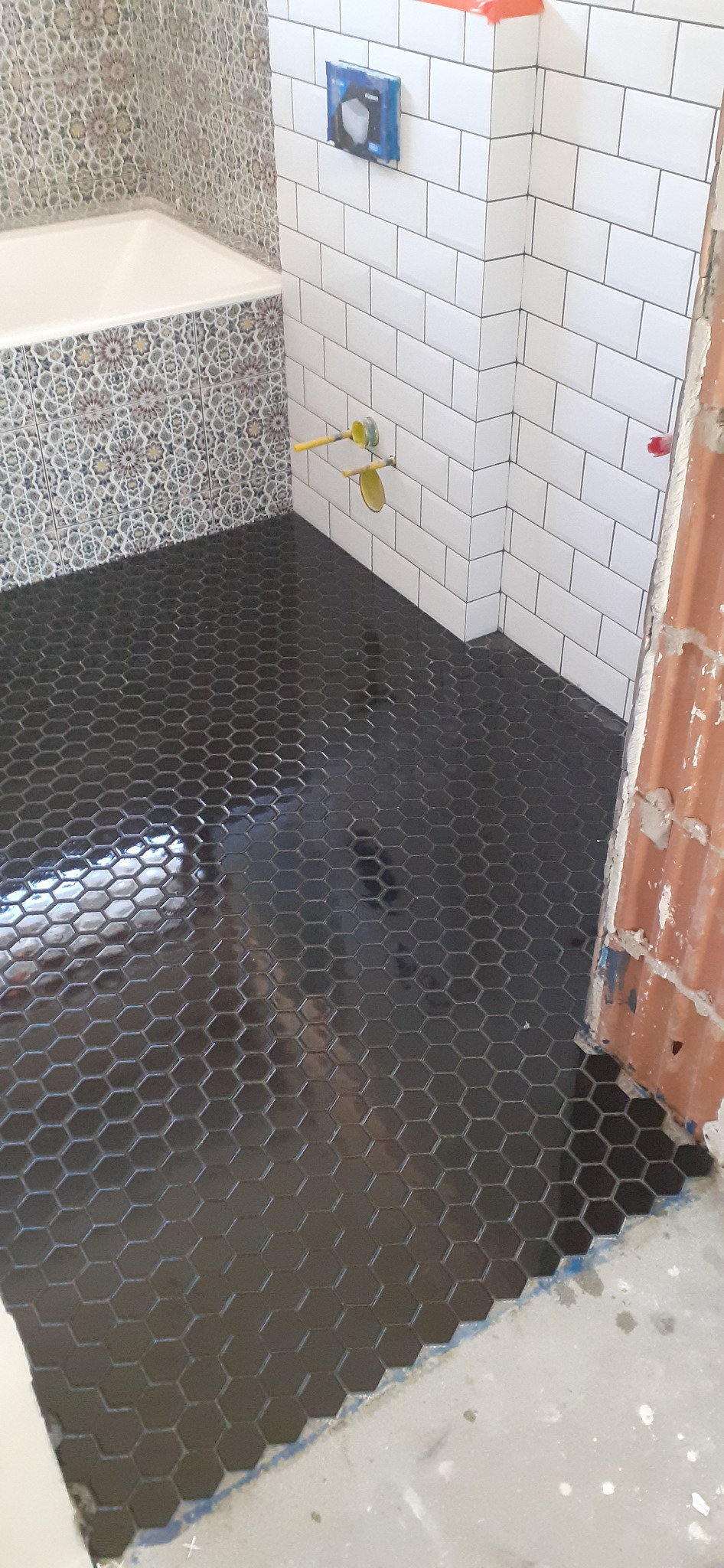 Czarne, heksagonalne płytki podłogowe w trakcie układania w łazience, widoczne białe płytki ścienne typu metro oraz fragment wanny obłożonej mozaiką wzorzystą.