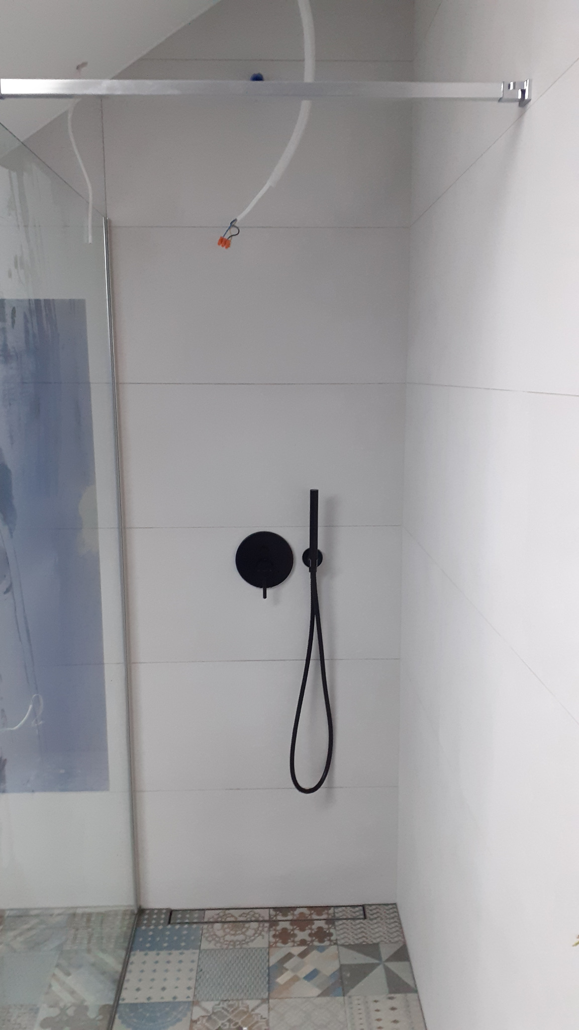 Nowoczesna kabina prysznicowa z czarną armaturą na tle białych płytek i wzorzystej podłogi, widoczne niedokończone podłączenia elektryczne pod sufitem.