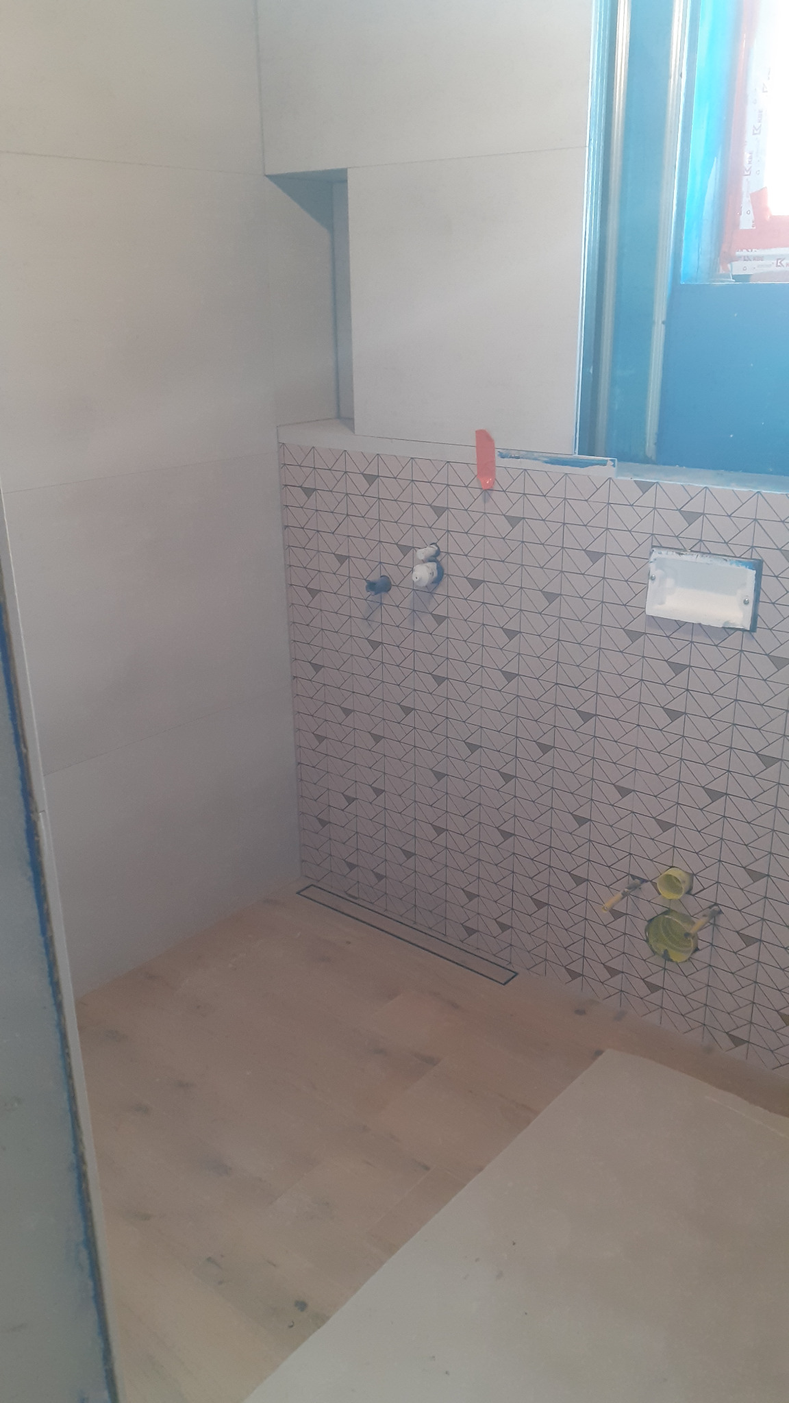 Remont łazienki w trakcie: ściany z geometrycznymi płytkami, zamontowany odpływ liniowy, przygotowane przyłącza wodne, widoczne elementy instalacji podtynkowej.