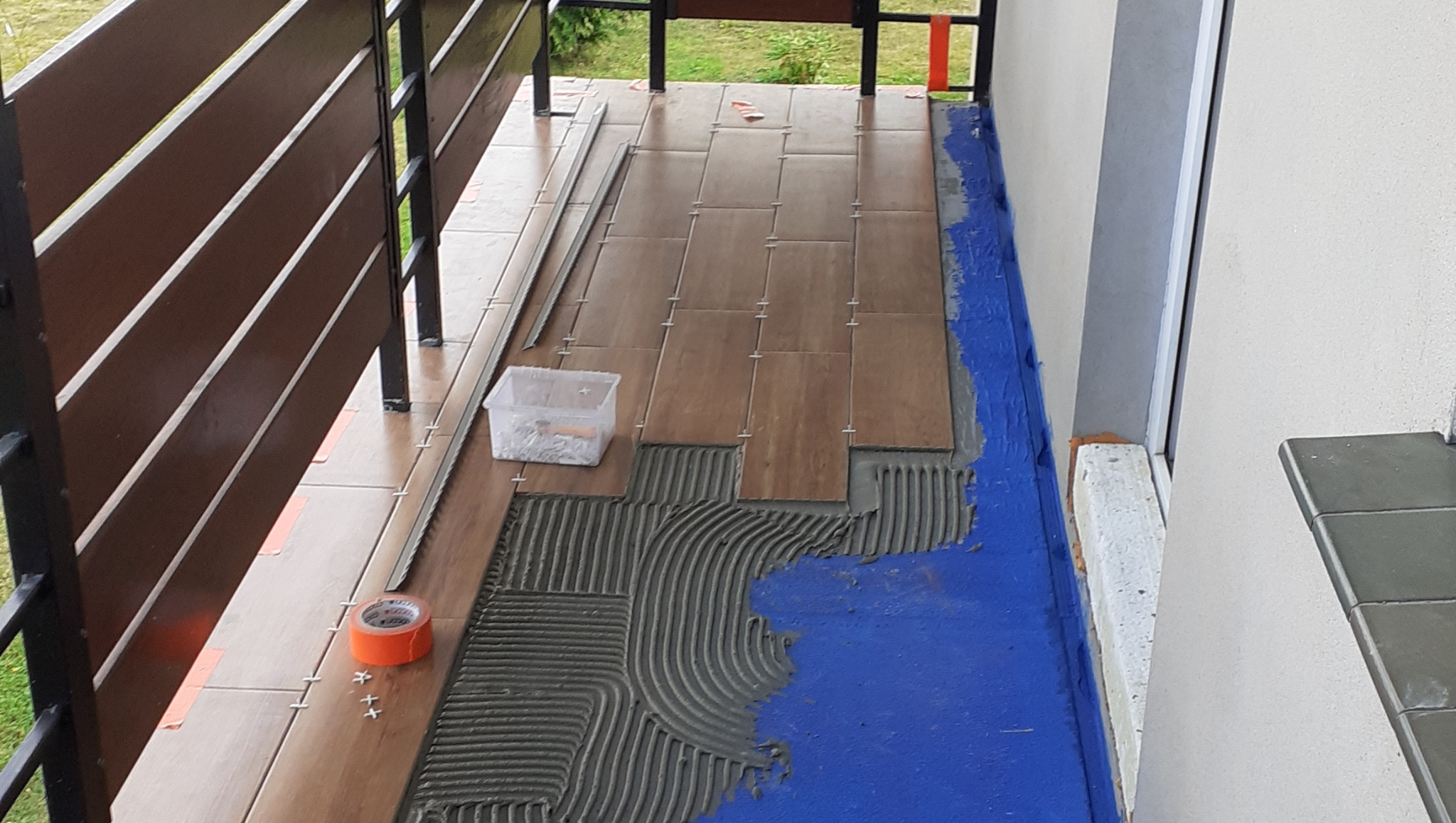 Układanie brązowych płytek na balkonie, widoczny klej, poziomica, krzyżyki dystansowe i taśma malarska.