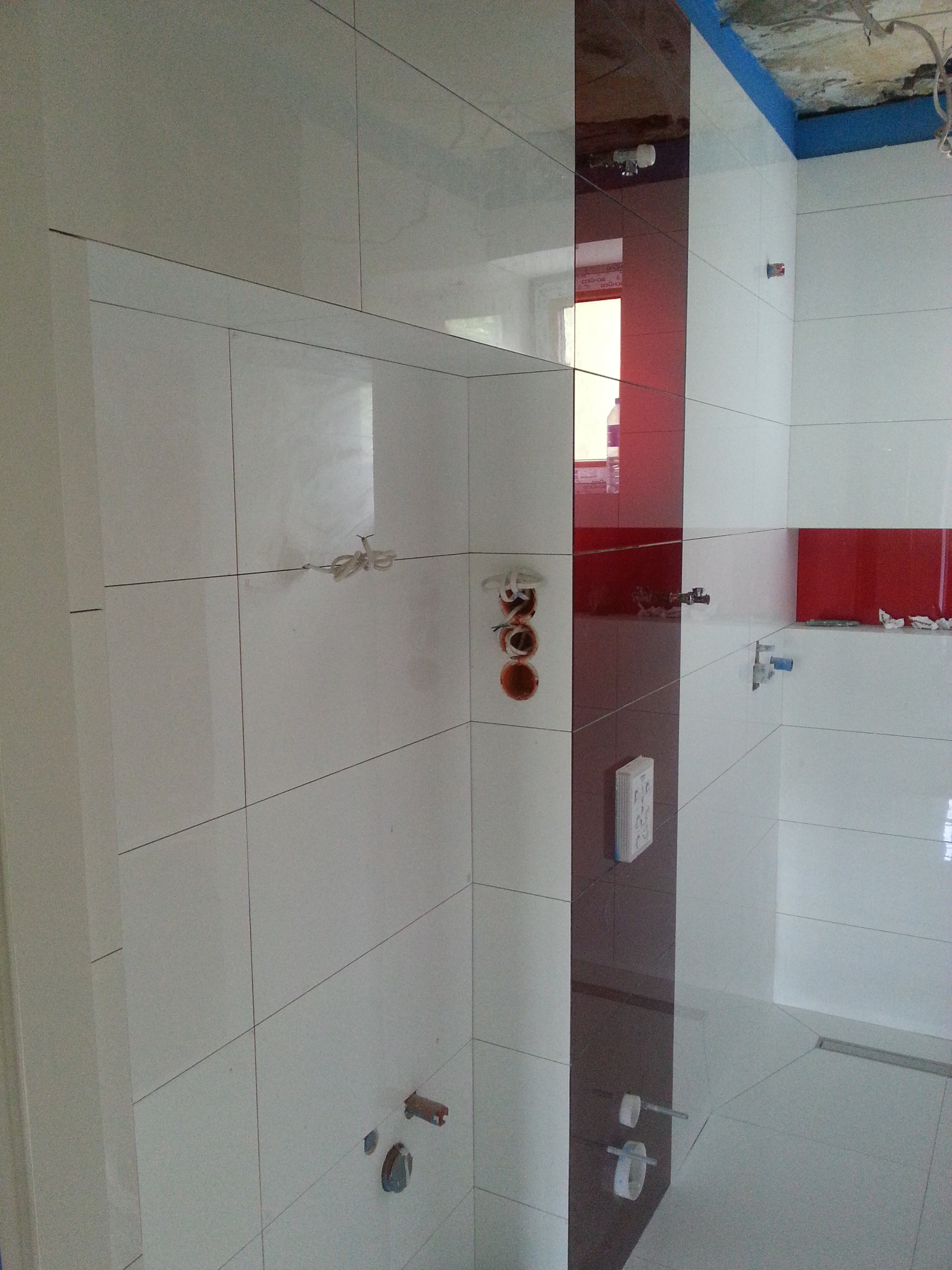 Łazienka w trakcie wykańczania: białe płytki częściowo ułożone, z akcentami w postaci pionowego pasa z ciemnoczerwonych płytek oraz poziomych pasów, widoczne otwory na instalacje, sufit...