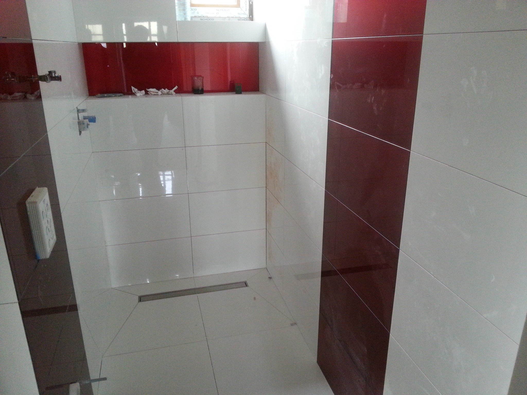 Wykończona kabina prysznicowa z biało-czerwonymi płytkami, odpływem liniowym w podłodze i półką z czerwonym szkłem.