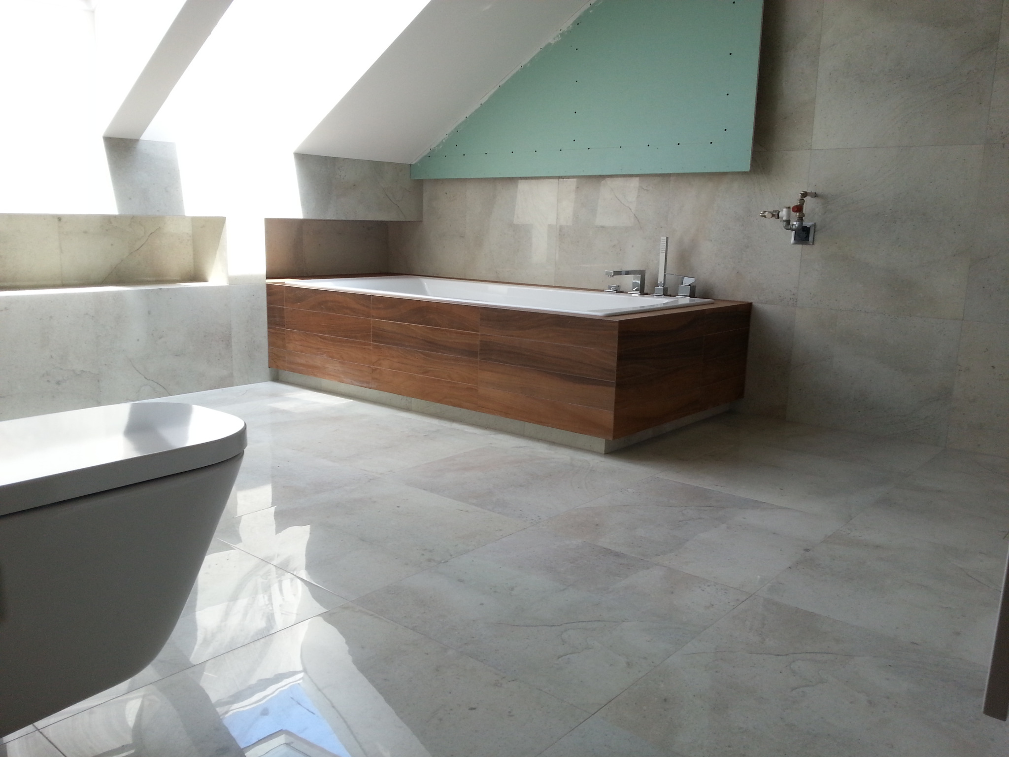 Wnętrze łazienki w trakcie prac wykończeniowych: wanna obudowana drewnopodobnymi płytkami, fragment ściany z płyt karton-gips, beżowe płytki podłogowe i ścienne, widoczna toaleta.