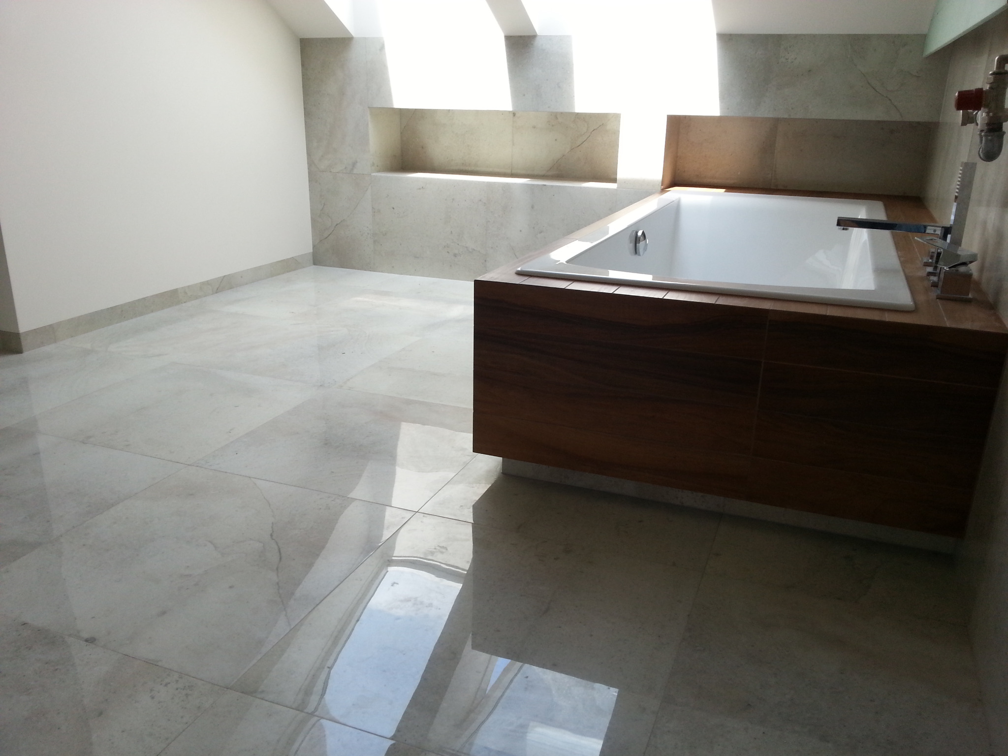 Nowoczesna łazienka z wolnostojącą wanną obudowaną drewnem, jasne płytki podłogowe i ścienne, okno dachowe wpuszczające naturalne światło.