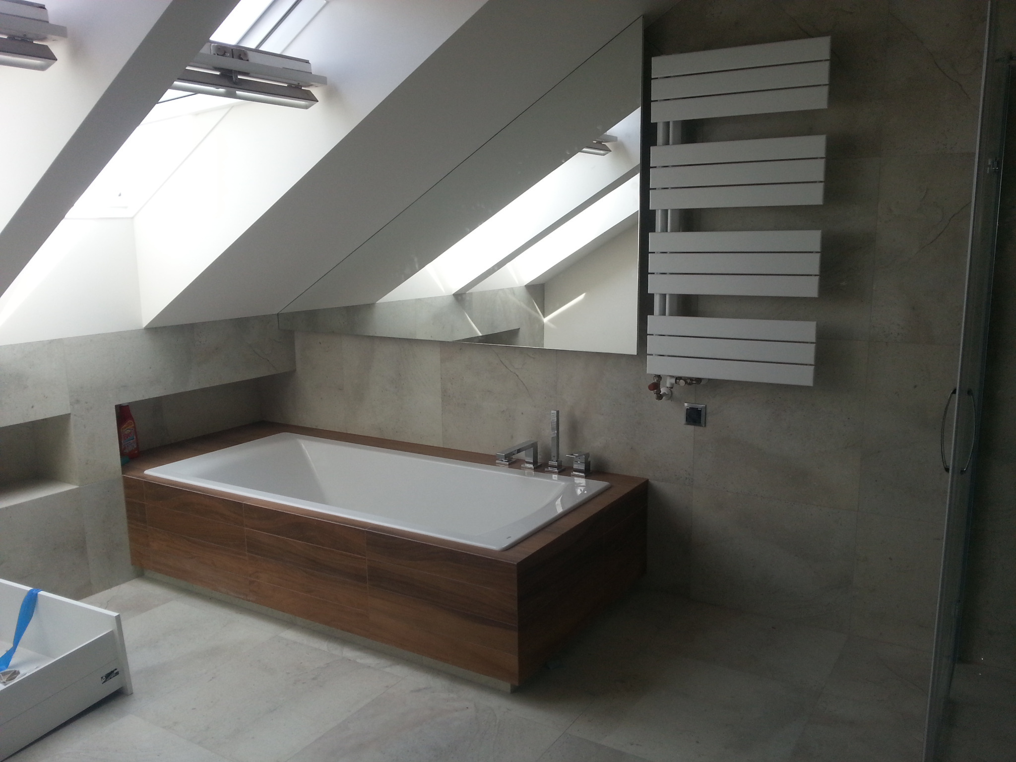 Nowoczesna łazienka na poddaszu ze skośnym sufitem, obudowana wanna z drewnopodobną okładziną, grzejnik drabinkowy, marmurowe płytki i okno dachowe.