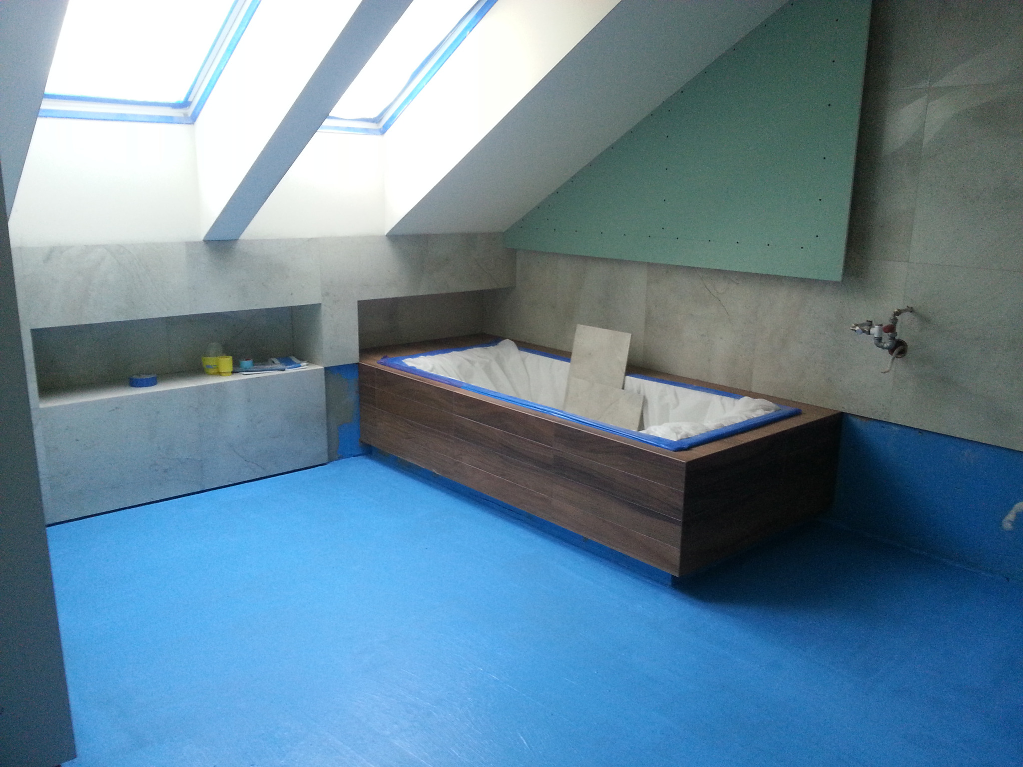 Łazienka w trakcie remontu: obudowana wanna z drewnopodobną okleiną, zabezpieczona folią, niebieska folia ochronna na podłodze, sufit skośny z oknem dachowym, ściana częściowo wyłożona płytkami...