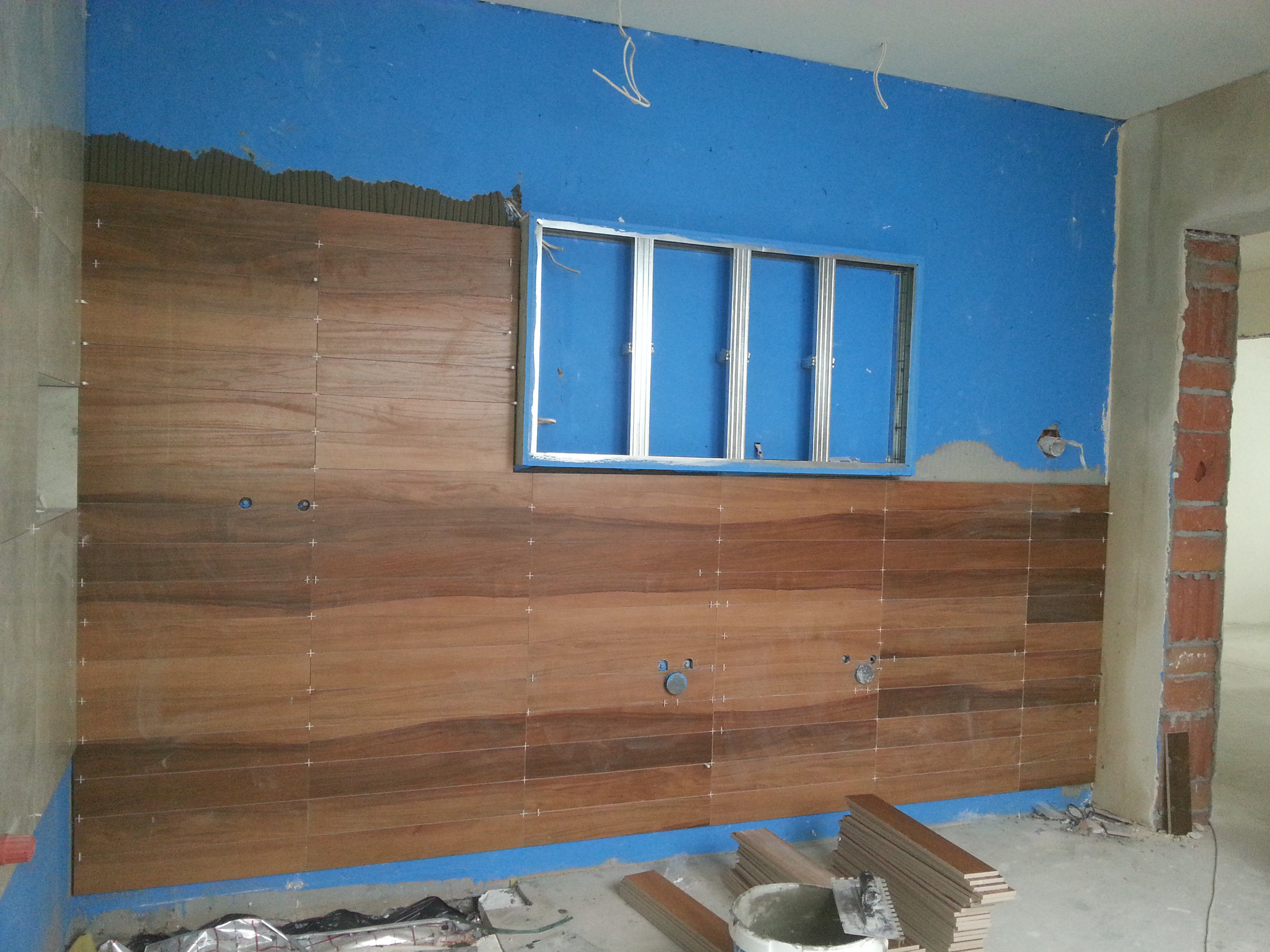 Ściana łazienki w trakcie wykańczania: ułożone brązowe płytki imitujące drewno z krzyżykami dystansowymi, niebieska ściana powyżej z metalową ramą okna, widoczne puszki elektryczne, wiadro i płytki...