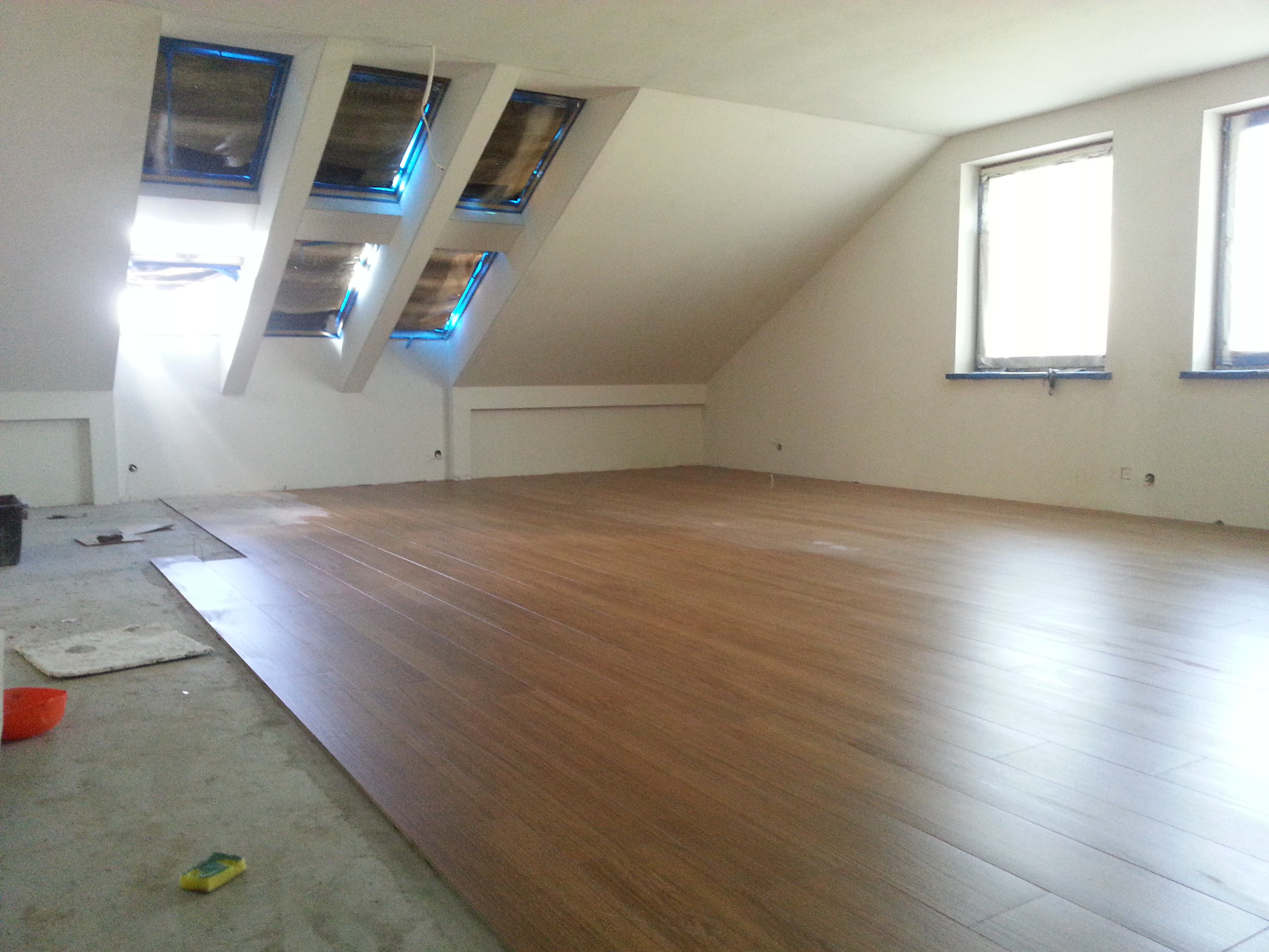 Układanie paneli podłogowych w jasnym, skośnym pokoju na poddaszu z oknami dachowymi i standardowymi oknami; widoczne przygotowane podłoże i narzędzia.