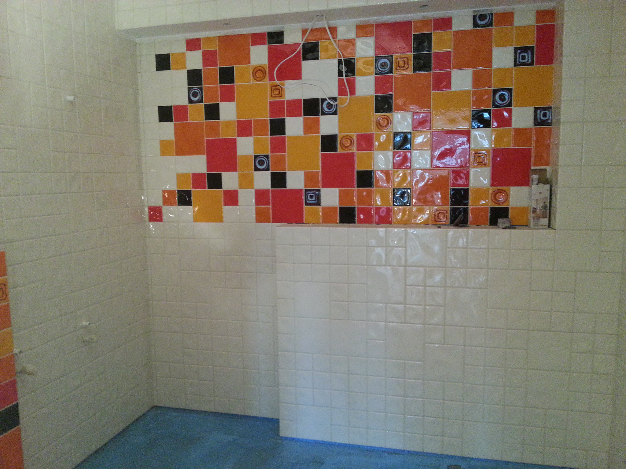 Ściany łazienki wyłożone białymi płytkami, z wnęką ozdobioną kolorowymi płytkami w odcieniach czerwieni, pomarańczy, czerni i bieli, widoczne przewody elektryczne.