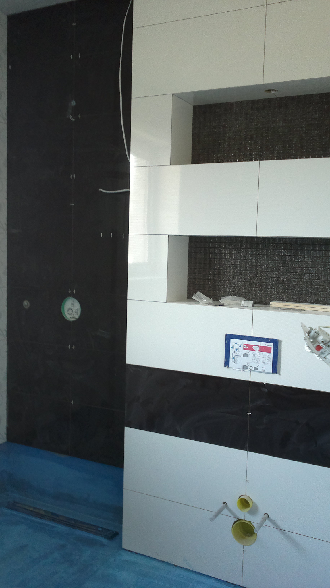 Układanie czarno-białych płytek w łazience w Zduńskiej Woli. Widoczne rury, poziomica i instrukcja montażu.